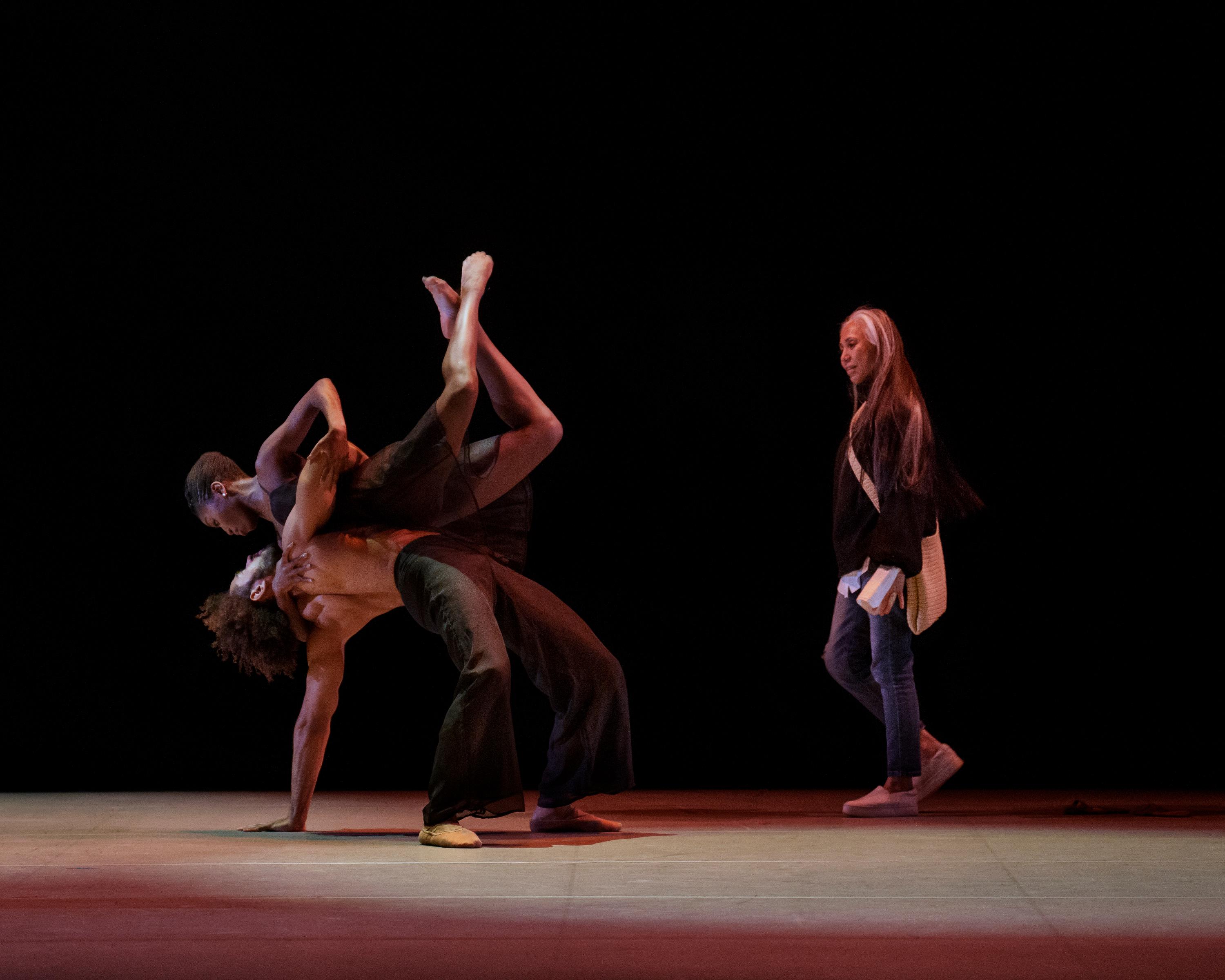 Two dancers rehearsing on stage while choreographer Elizabeth Roxas-Dobrish observes and gives notes from the side