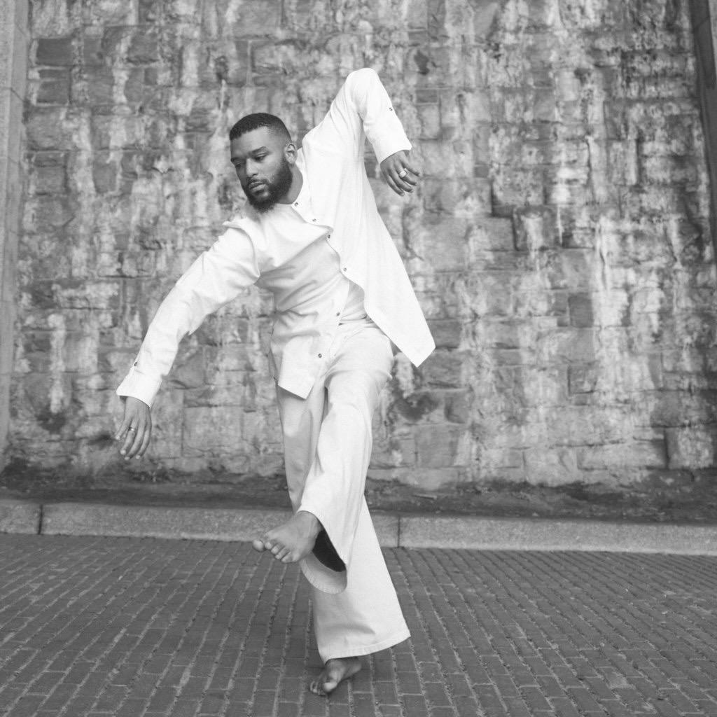 A black and white photo of a Black male contemporary dancer with short hair and a short beard dressed in all white long sleeve pants and a long sleeve button-up shirt
