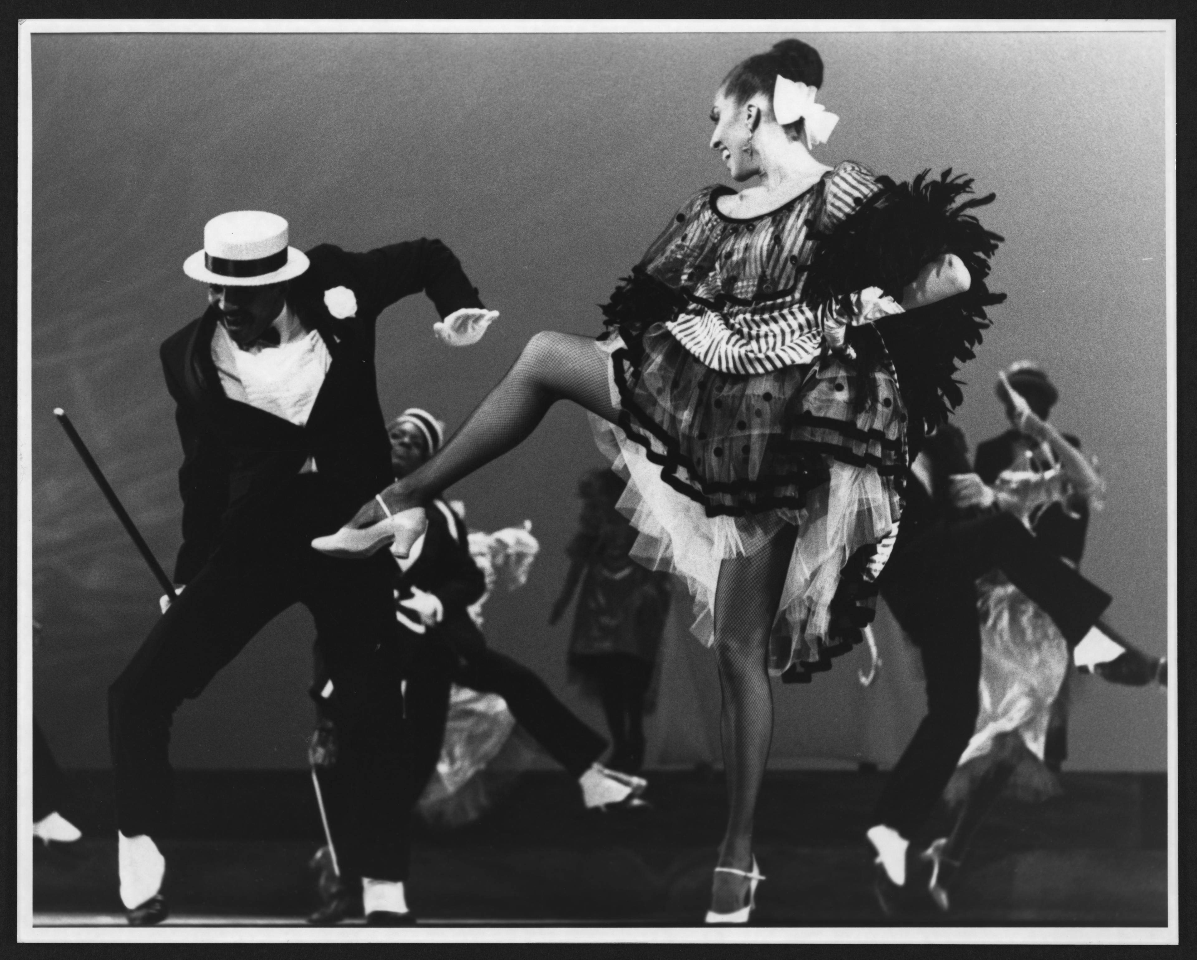 Black and white photo of two dancers. One is in a top hat and one is in a dress with their leg lifted to the side.