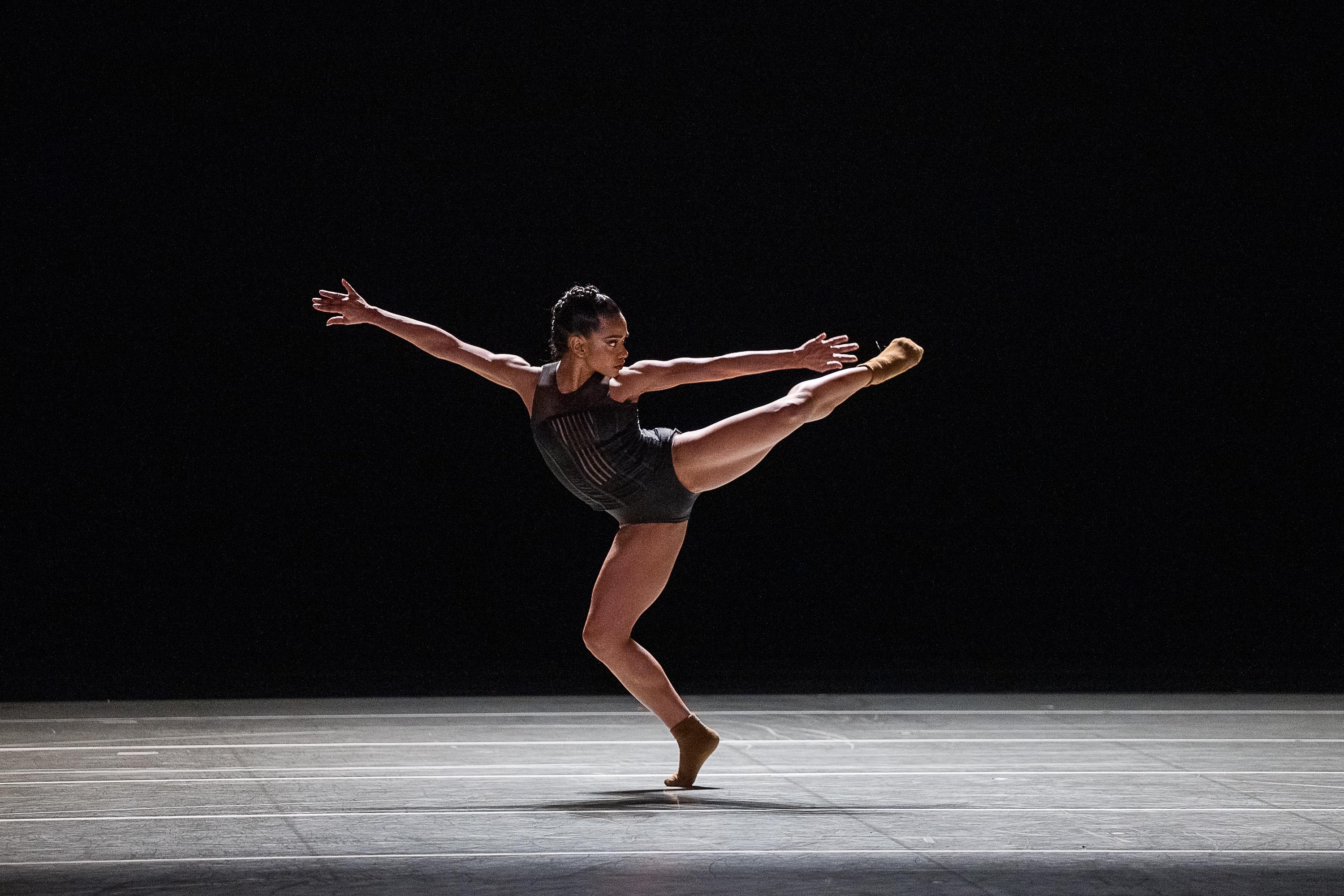 Female dancer wearing black in front of a black background with arms and leg extended