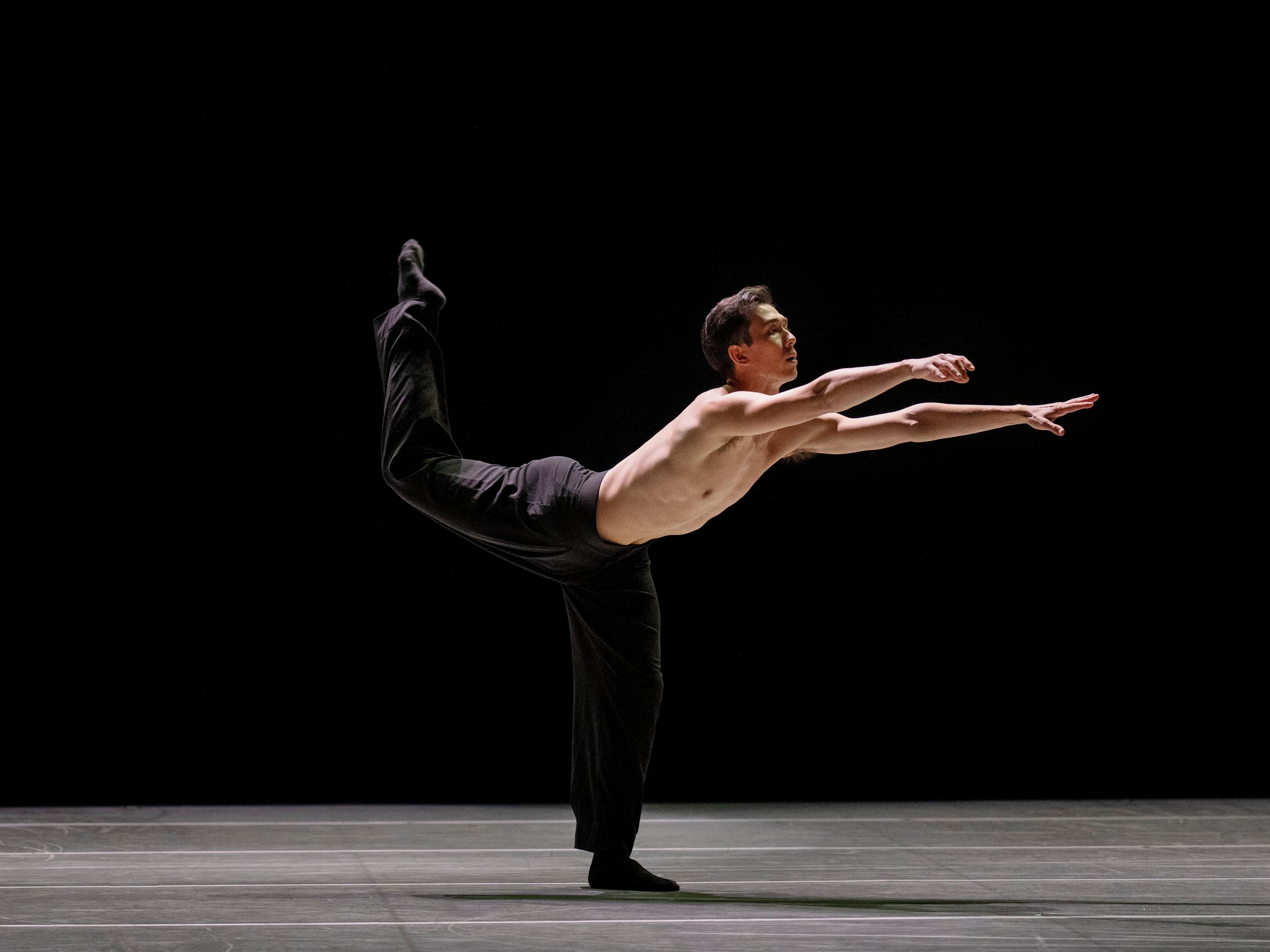 A dancer in black pants with one leg up behind them and arms reaching forward
