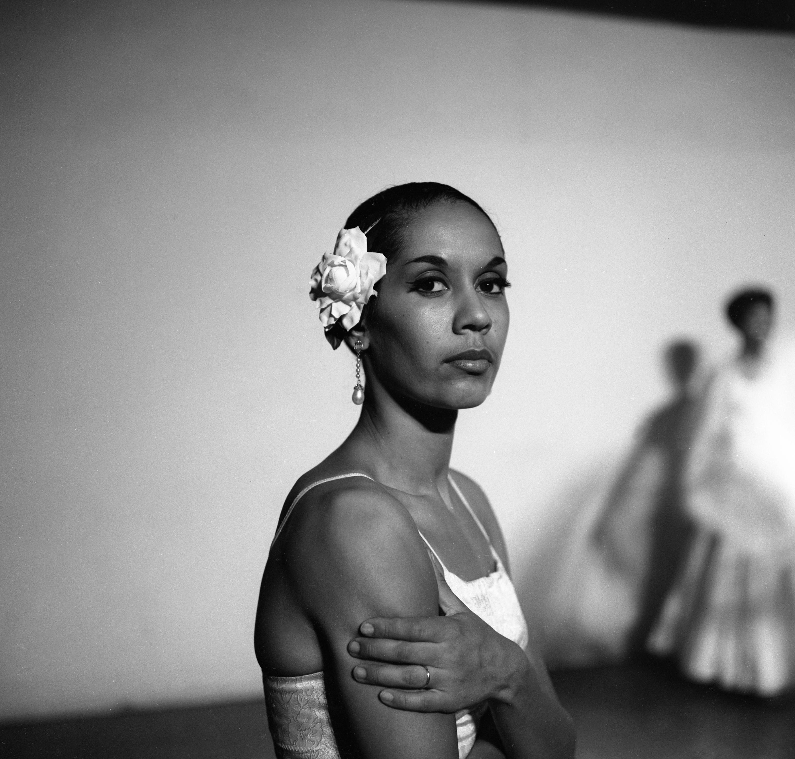 Carmen de Lavallade in black and white wearing a white flower in her hair and white dress looking at the camera holding her arm