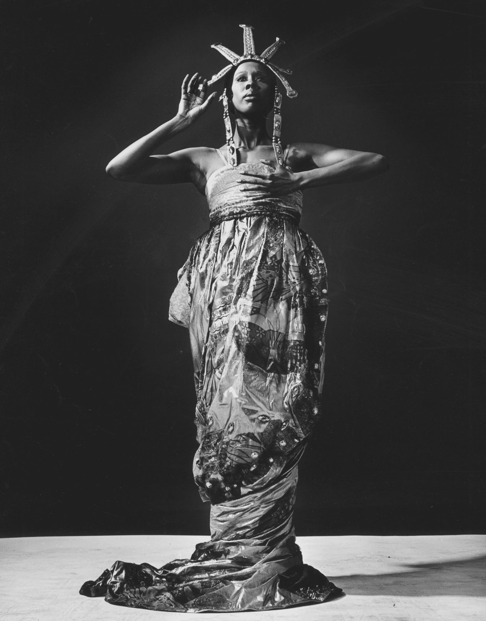 A dancer stands with one arm bent and hand raised to the head, wearing an elaborate headdress and a strapless, flowing gown. The background is dark and the floor is visible.