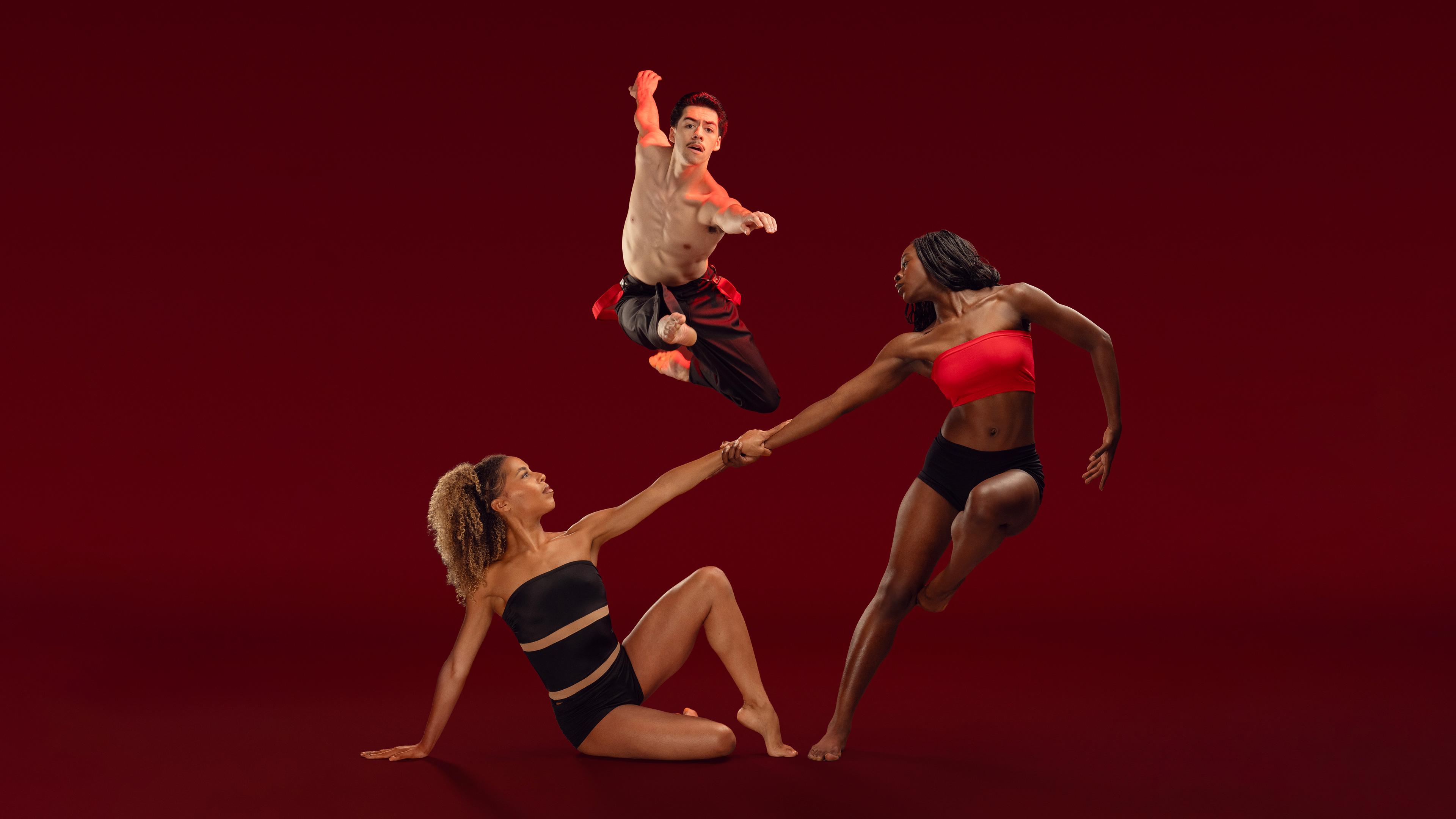 3 Ailey II dancers, 2 holding hands and leaning away from each other and the 3rd jumping behind