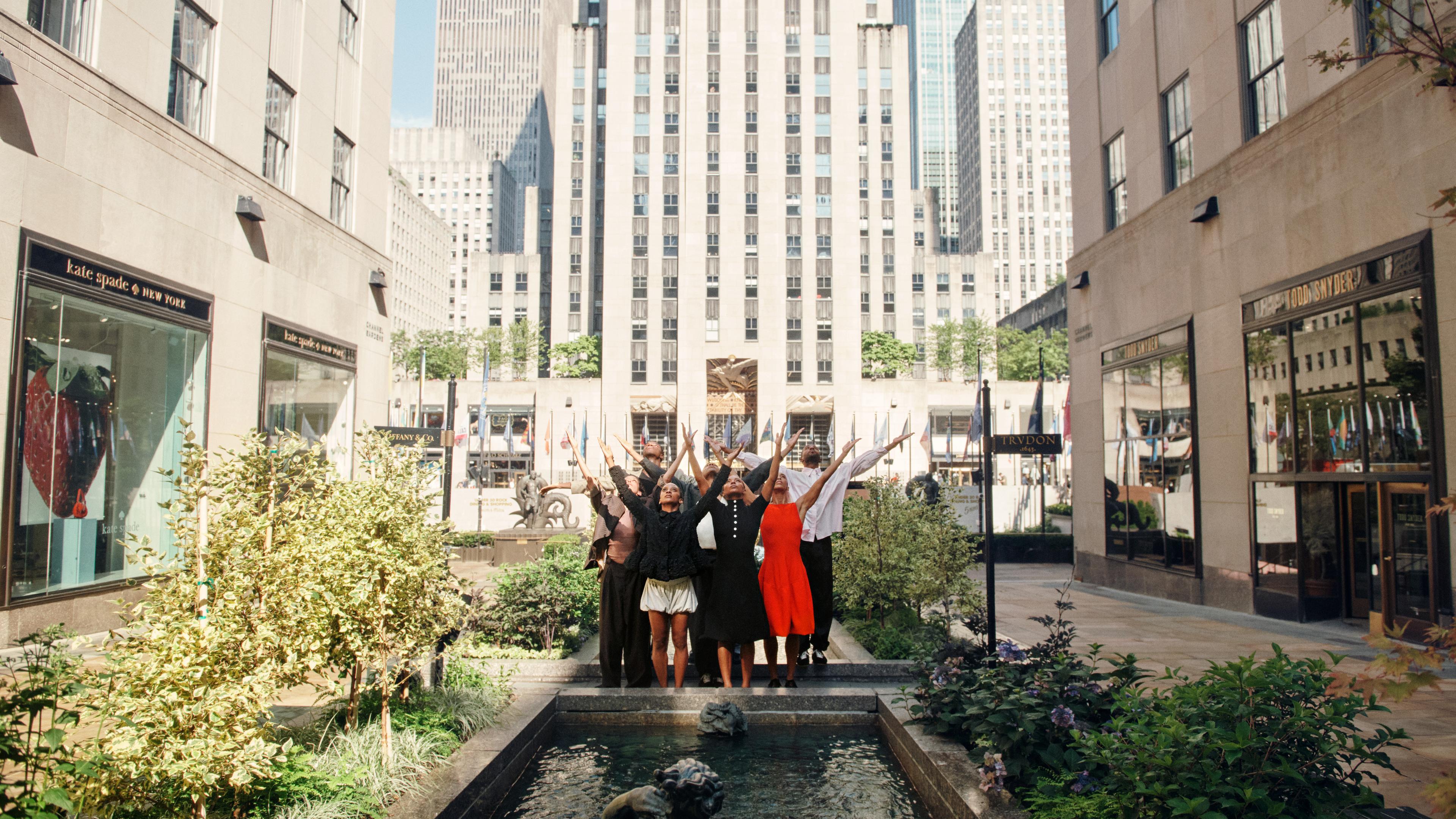 Ailey dancers in Rockefeller Center plaza with their arms upward toward the sky