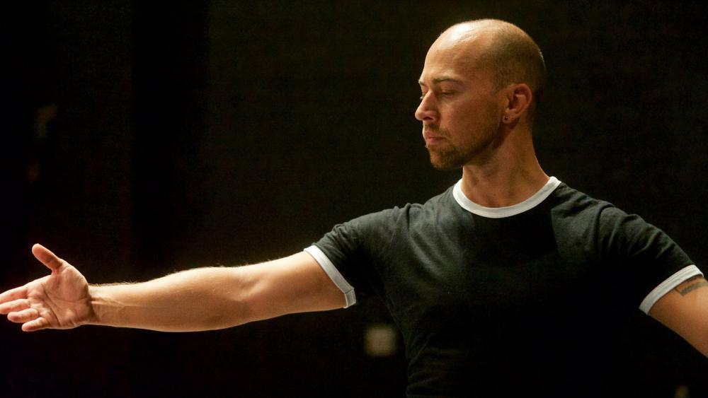 A man looks to the left in a black tee on a black background. His arms are outstretched.