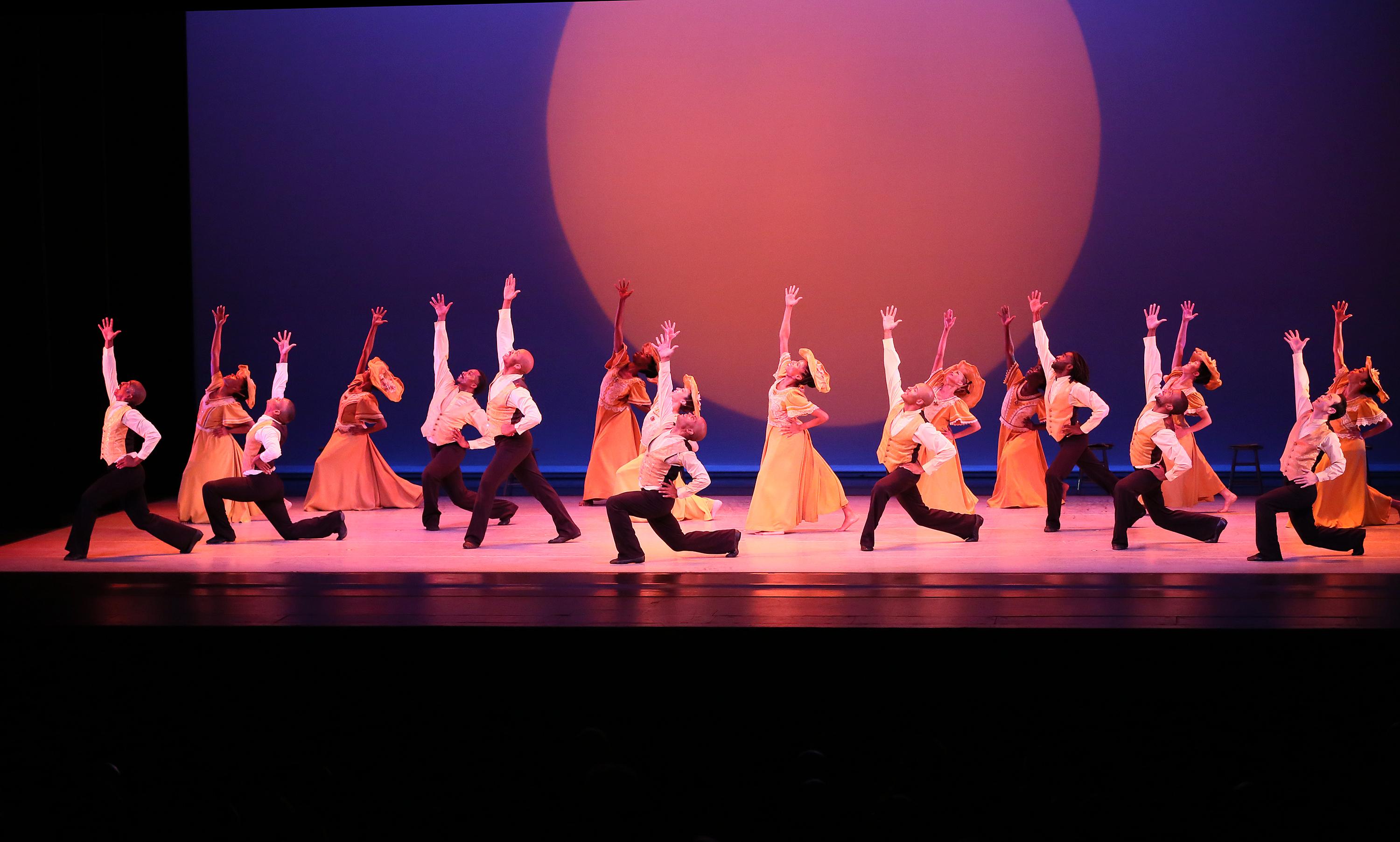 An ensemble of dancers in yellow costumes looking upward toward 1 arm with 1 knee bent