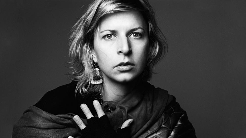 A woman stares at the camera with a scarf. Her hand is on her chest.