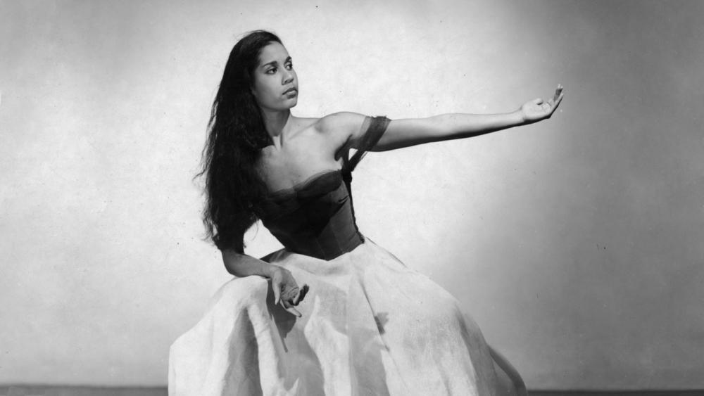 A woman in black and white sits with her arm outstretched. She is in a black top and a white skirt.
