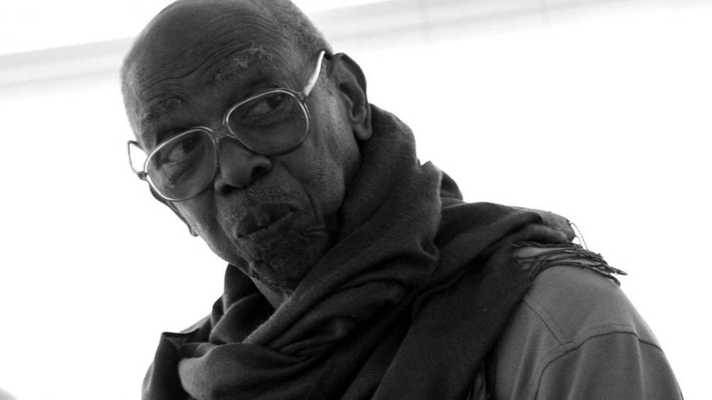 A black man with wire glasses looks off to the right. He has a scarf and a tee on.