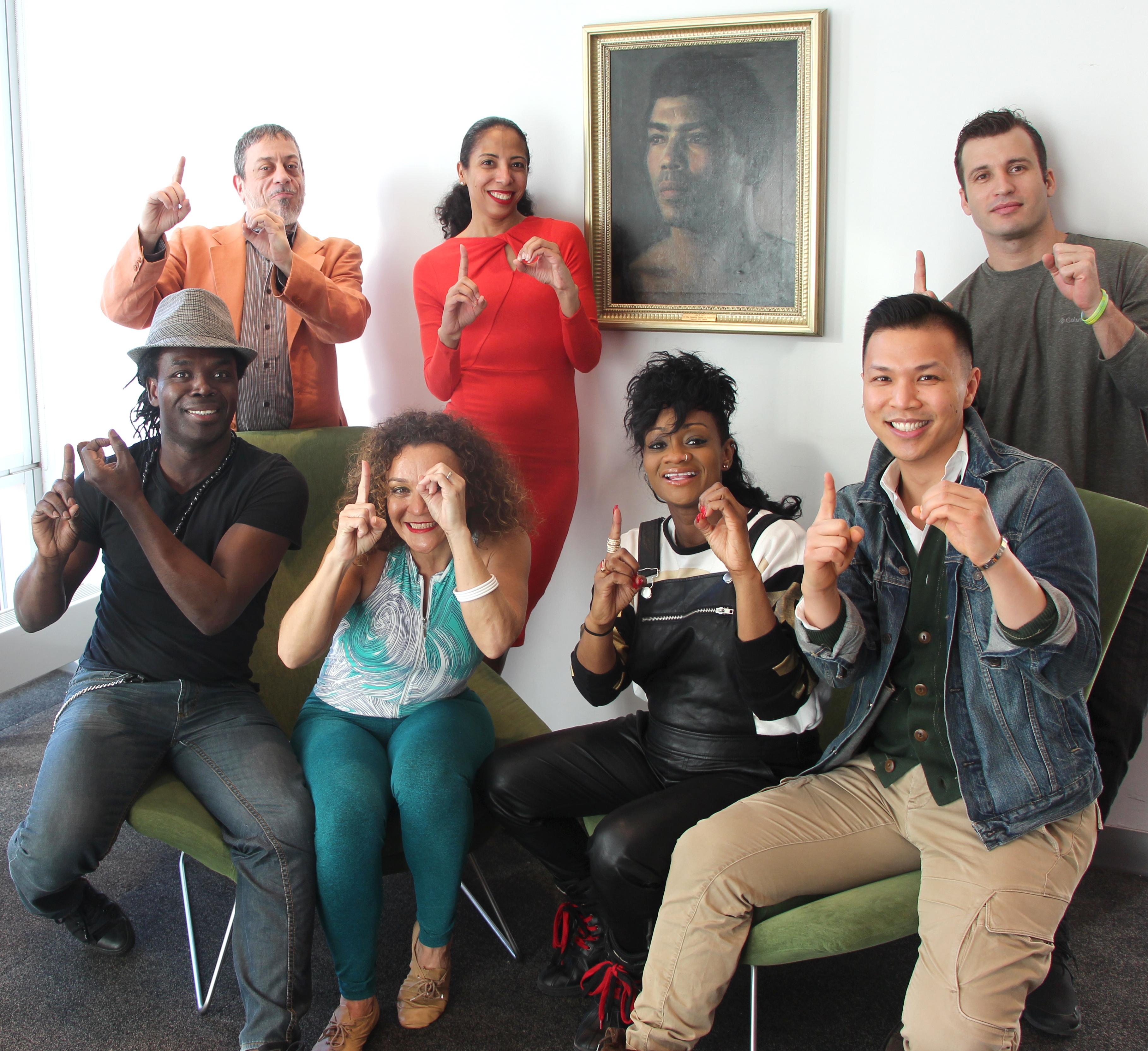 A color photo of the same group of seven people from the previous image, now all holding up one finger each. They are still posing in the same room with the portrait on the wall, smiling and displaying a gesture of unity.