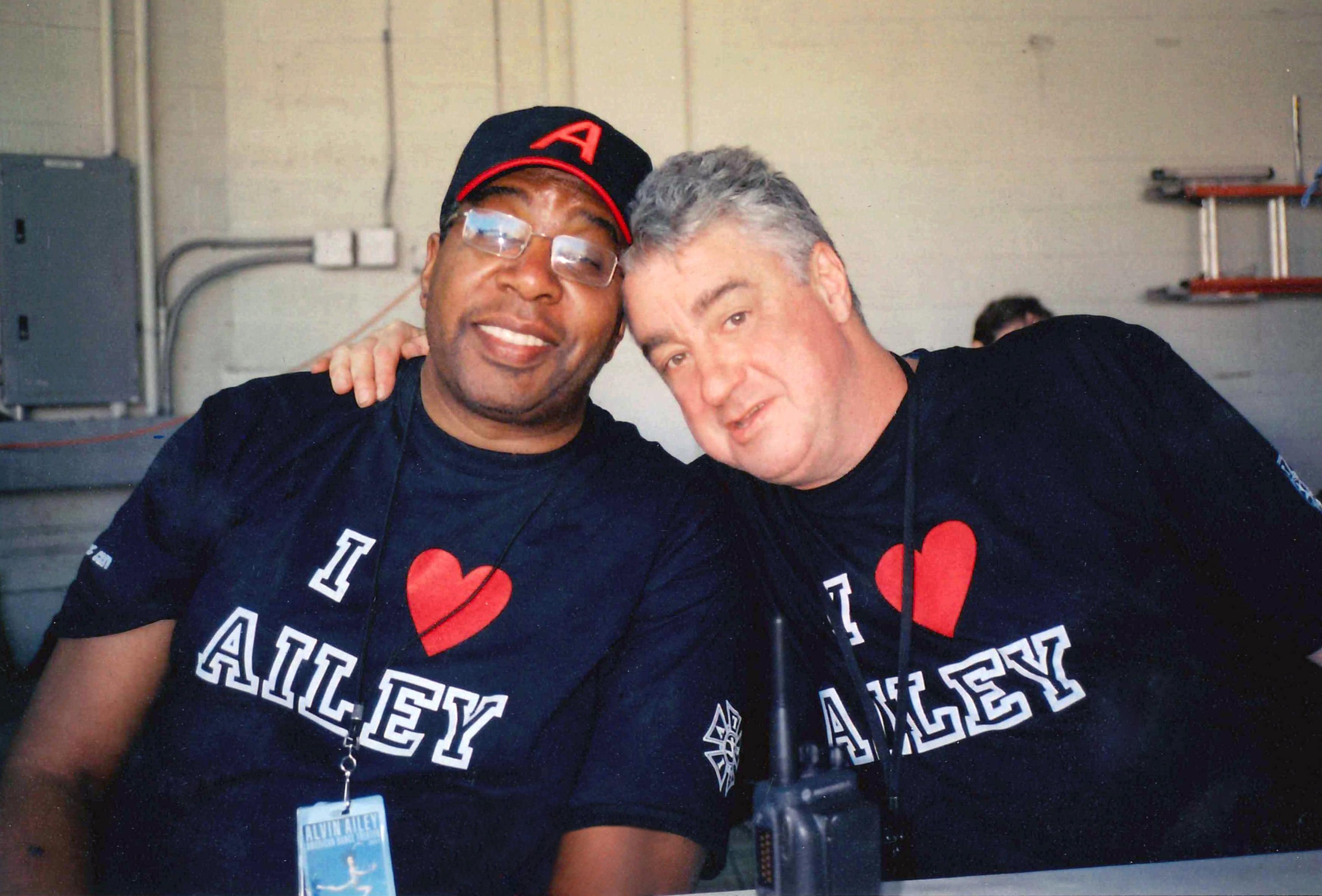 Two men are sitting hugging with blue shirts that say I <3 AILEY on them