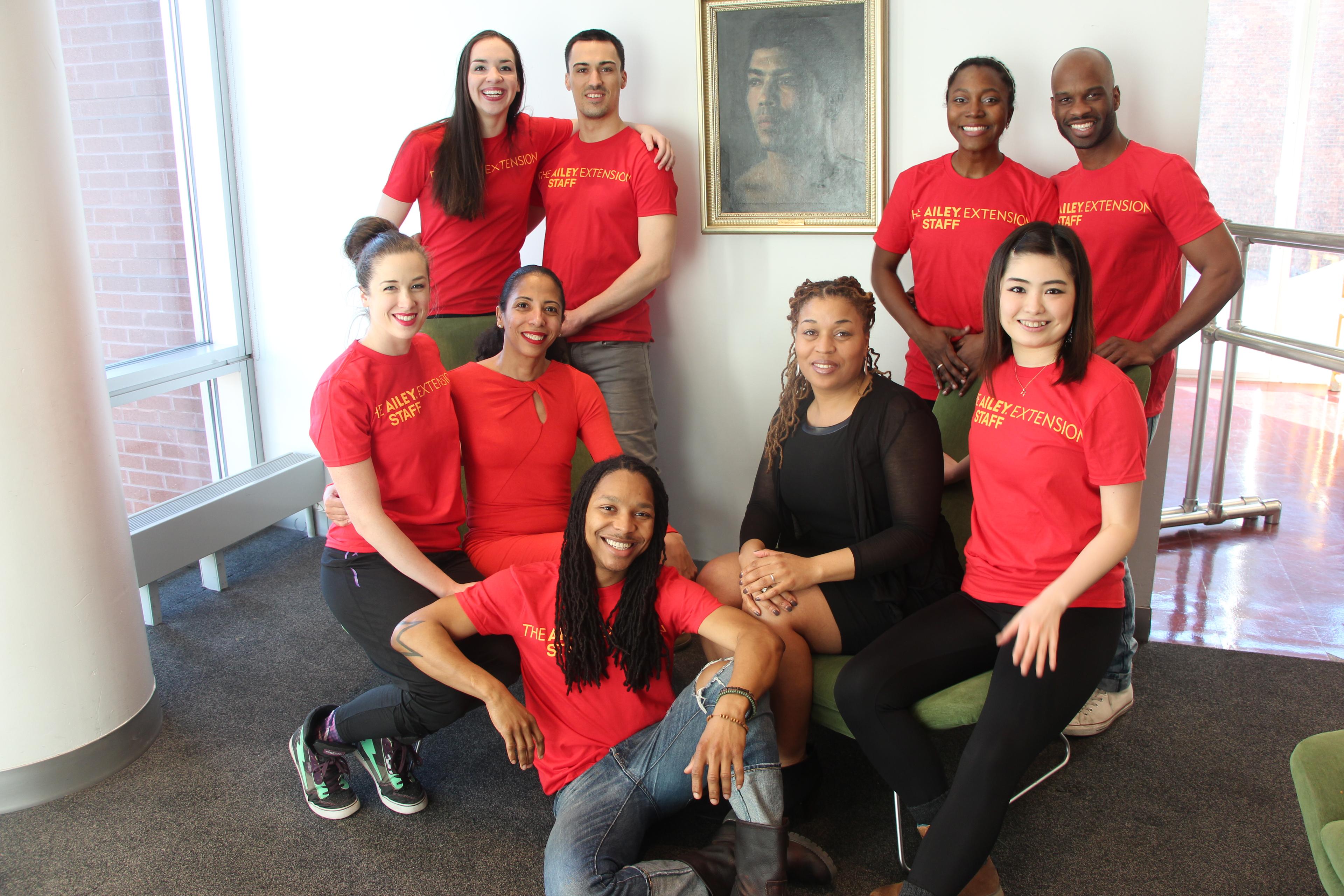 A color photo of a group of nine people, with eight wearing matching red Ailey Extension shirts and one person in black, posing together in a room. They are smiling and sitting or standing around a seated woman, with a portrait on the wall in the background.