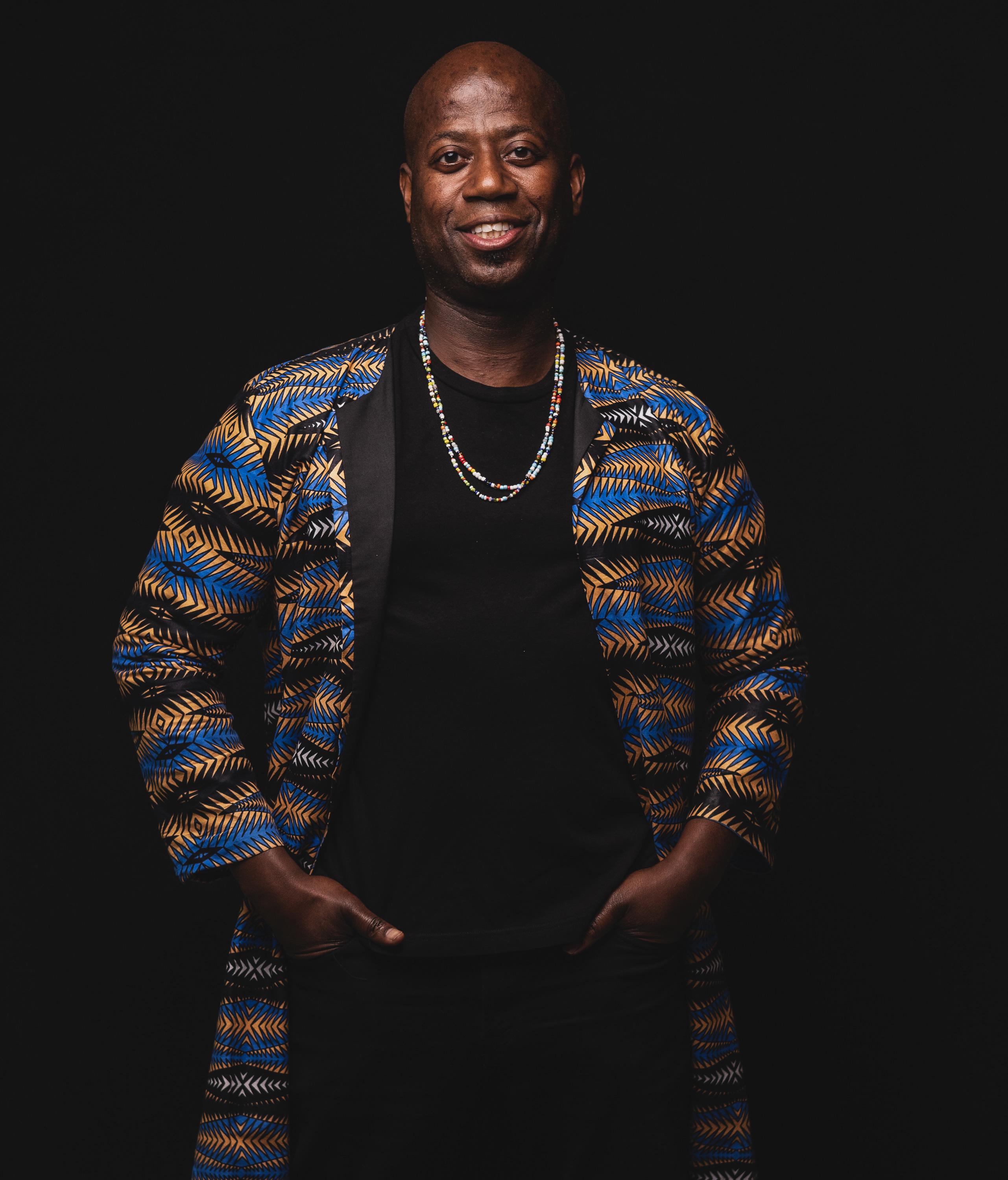 A headshot of a man against a black backdrop wearing striped cardigan, black shirt, and necklace