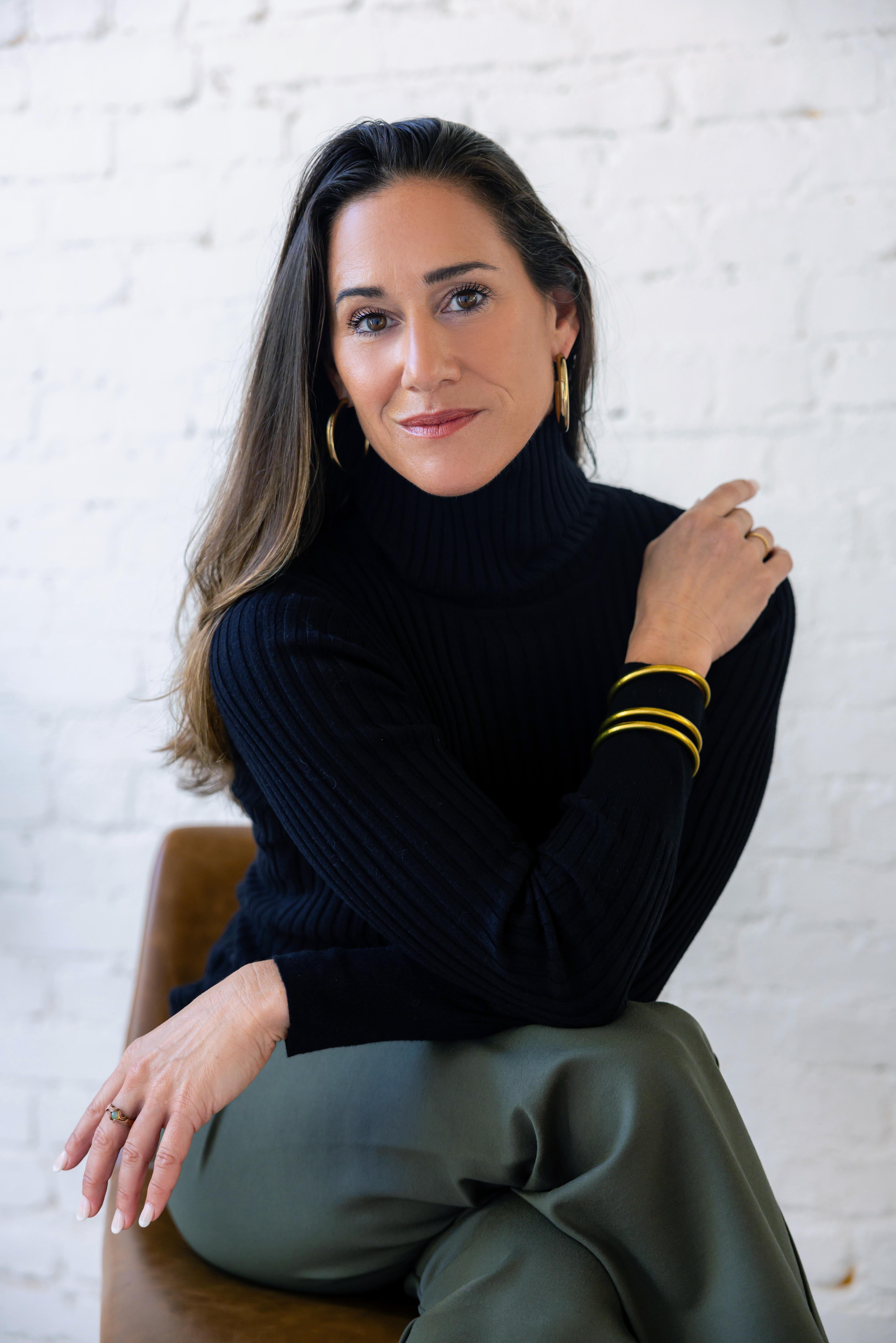 A headshot of Maija Garcia with long brown hair, wearing a black turtleneck, green pants, and gold jewelry