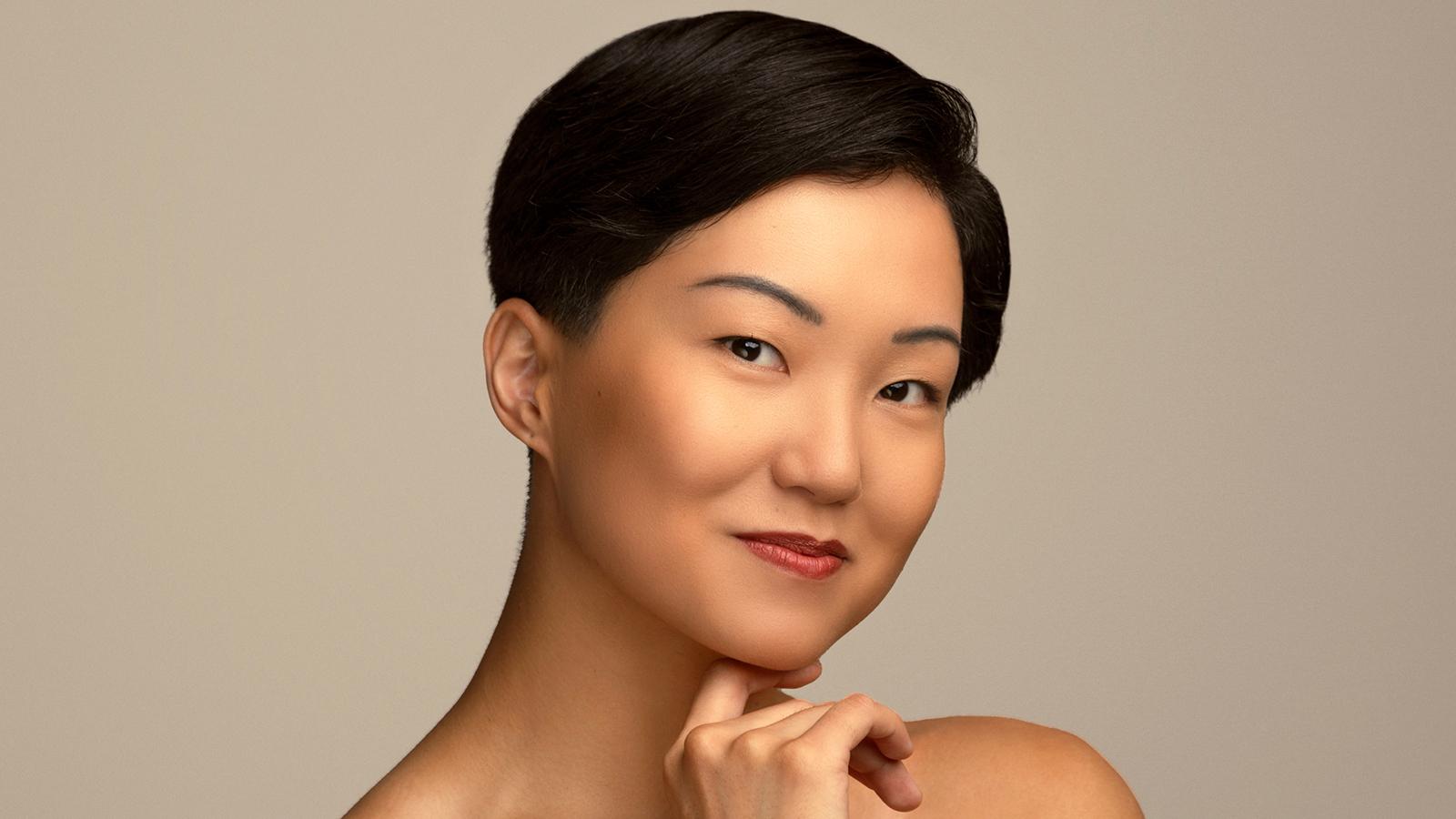A headshot of Miranda Quinn, a dancer with short dark hair, smirking with her hand under her chin