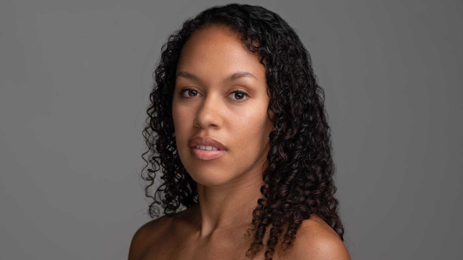 Photograph of a woman with medium-dark skin and long curly black hair, looking directly at the camera with a neutral expression. The background is plain gray.